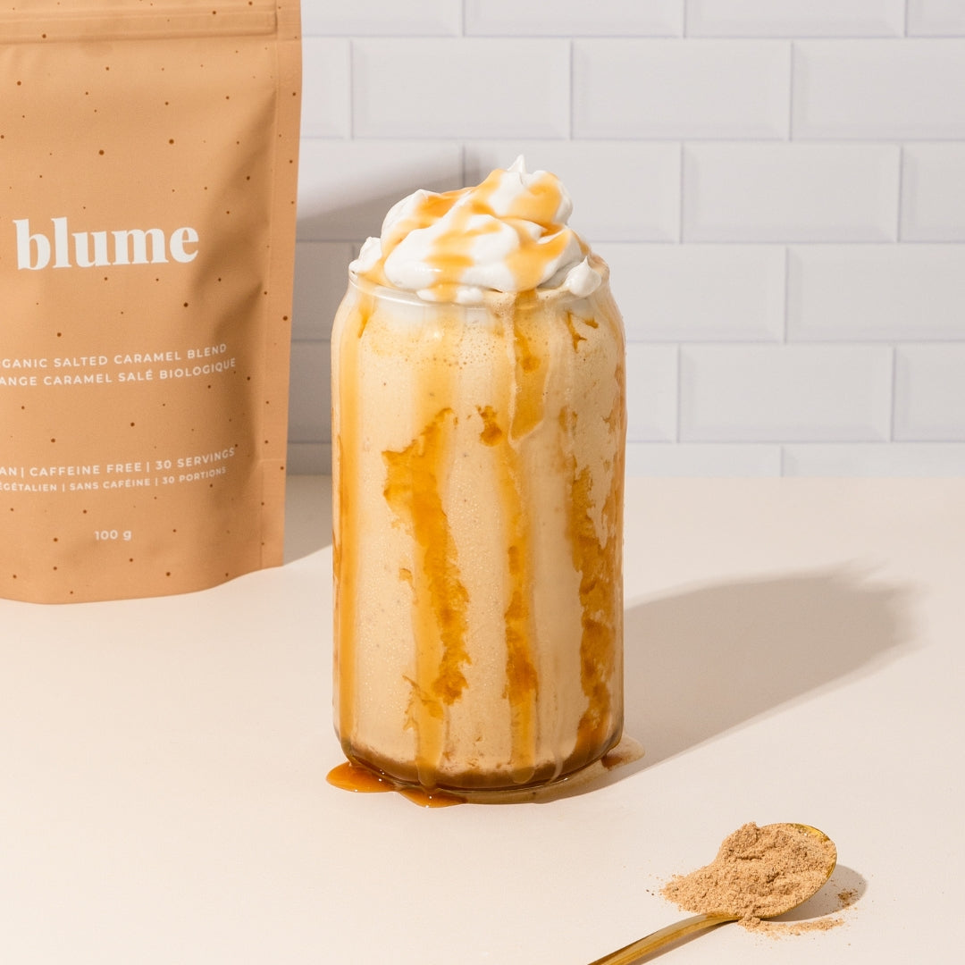 A creamy drink topped with whipped cream and caramel drizzle in a glass jar on a white table. Beside it is a golden spoon with powder and a bag labeled blume for organic salted caramel blend against a tiled backdrop.